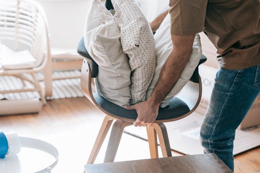 A man packing throw pillows on a contemporary chair indoors, suggesting moving or organizing.